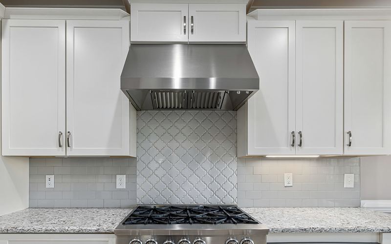 Stainless range hood over a gas range, with white cabinets and tile backsplash